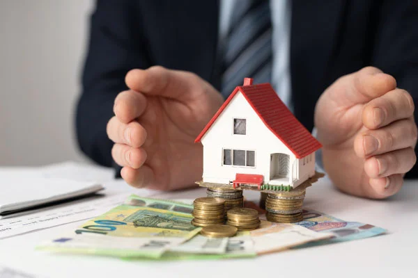 Model house standing on some coins and bills and male hands surrounding it | 5 money tips to share with your borrowers | DG Pinnacle TPO - Miami Wholesale Mortgage Funding Lender